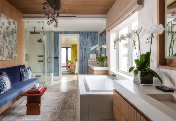 A bright, refined bathroom with a freestanding white bathtub beside a broad vanity and integrated basin. The room includes a glass-enclosed shower, timber cabinetry and ceiling, blue patterned wall panels, a built-in blue upholstered bench, large windows, and orchids adding a natural decorative element. A doorway leads toward another interior space.