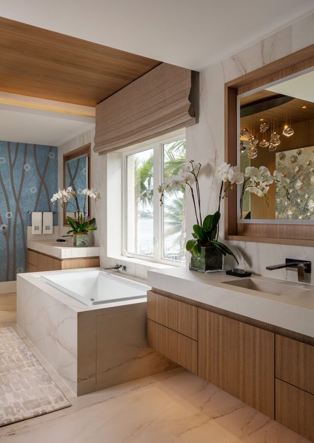 A sophisticated contemporary bathroom with a freestanding-style rectangular bathtub set into a marble-clad surround, a long floating timber vanity with pale stone benchtop and an undermount basin, and a large framed mirror. Warm timber ceiling lining and cabinetry contrast with pale veined stone surfaces. A sculptural pendant cluster, orchids, patterned wallcovering, and a window with tropical foliage add decorative detail.