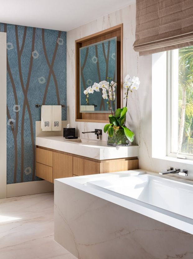 Bright, refined bathroom featuring a freestanding white bathtub in the foreground, a floating timber vanity with a white stone benchtop, wall-mounted tapware, a large timber-framed mirror, and a patterned blue feature wall with organic brown lines and geometric motifs. A woven Roman blind covers the window, with tropical greenery visible outside. The space combines warm timber, pale marble-look surfaces, and decorative patterned wallcovering.