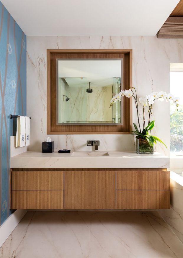 A bright, refined bathroom vanity area featuring a floating warm timber cabinet with integrated drawers, a pale stone countertop and undermount basin, and a large rectangular mirror framed in matching timber. The walls and floor are clad in white marble-look surfaces with subtle veining. A blue decorative wall panel appears at left, while an orchid and natural daylight add softness. The mirror reveals a glass shower area behind the camera-facing vanity.