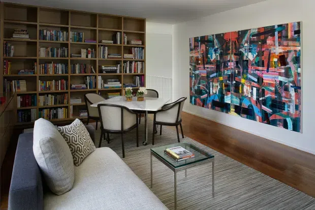A refined living and dining area with a large built-in timber bookcase, a light upholstered sofa, glass coffee table, round dining table with curved chairs, and a striking abstract artwork. The room has warm timber flooring, neutral walls, and a textured area rug, creating a sophisticated but inviting atmosphere.