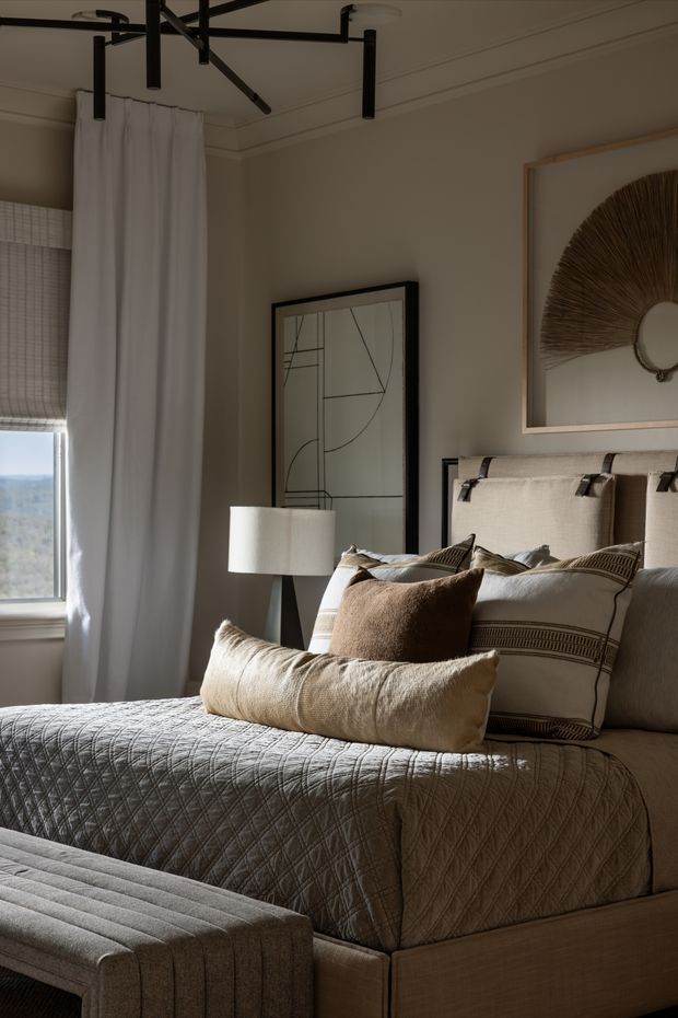 A refined, warmly lit bedroom with a neutral beige and taupe palette. The room features a textured upholstered bed with layered cream and brown cushions, a quilted bedspread, a matching upholstered bench, framed abstract artwork, a woven sunburst wall piece, white curtains, and a sculptural black ceiling light. A window provides natural daylight and an outdoor view.