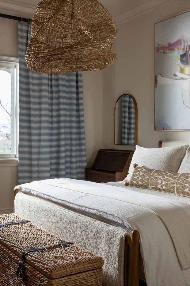 A softly lit, warm bedroom featuring a neatly made bed with layered white textured bedding, a patterned neutral cushion, a dark wood writing desk, an arched wall mirror, blue-and-white striped curtains, and a large woven rattan pendant light. A woven wicker trunk sits at the foot of the bed, while an abstract artwork adds colour to the neutral walls.