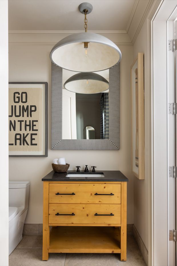 A compact, warmly styled bathroom with a honey-toned timber vanity, dark stone benchtop, undermount sink and black tapware. A large rectangular mirror with a bold black-and-white striped frame sits above the vanity, reflecting a matching pendant light. The room includes a white toilet, neutral painted walls, tiled flooring, white trim and a second framed artwork.