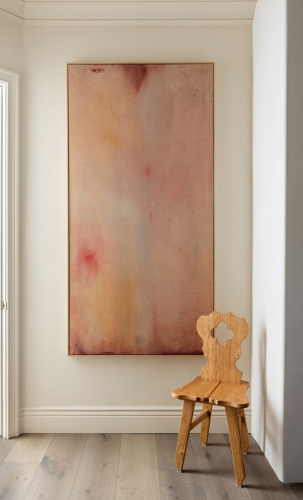 A softly lit, refined interior corner featuring a large abstract blush-toned artwork in a slim wood frame, mounted on a warm off-white paneled wall. A sculptural natural timber chair sits beneath the artwork, with pale timber flooring, white trim, and a bright doorway or opening at the side.