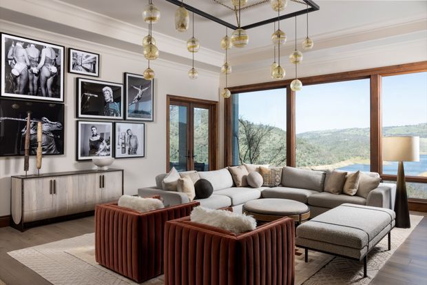 A refined living room with a large sectional sofa, ottoman, upholstered lounge chairs, and a textured area rug. The room features extensive timber-framed glazing overlooking a hilly landscape, a gallery wall of black-and-white artwork, a light timber sideboard, decorative wall moulding, and a striking black ceiling-mounted grid with numerous suspended glass pendant lights.