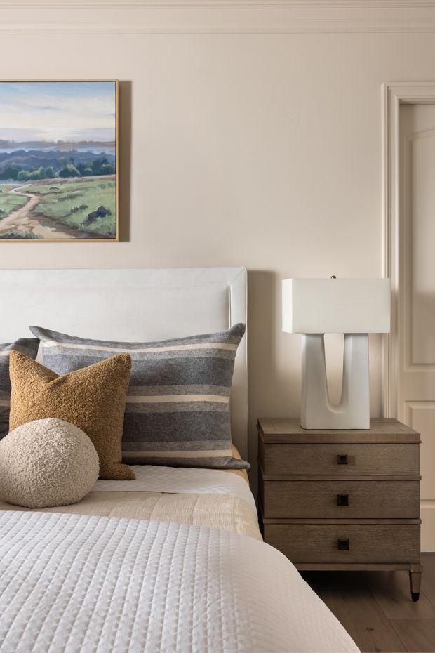 A softly styled bedroom corner with a white upholstered bed, layered neutral and grey striped bedding, textured mustard and cream cushions, and a wood three-drawer bedside table. A sculptural white table lamp sits beside the bed, while a framed landscape artwork and traditional moulded doorway add decorative detail. The palette is warm, neutral, and refined.