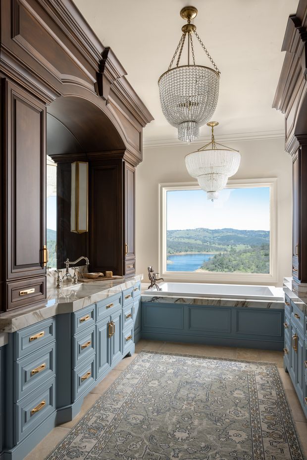 A luxurious traditional bathroom with a freestanding-style built-in bathtub beneath a large picture window overlooking a lake and rolling hills. The room features extensive dark timber cabinetry and moulded panelled joinery, pale blue-grey vanity cabinetry with brass hardware, white marble benchtops, ornate crystal pendant chandeliers, and a large patterned rug.