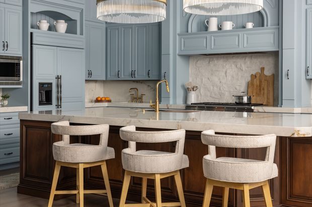 A refined, generously sized kitchen with blue-grey panelled cabinetry, dark timber island joinery, a pale stone benchtop, brass tapware, and a textured natural-stone splashback. Three upholstered bar stools sit at the island beneath oversized pendant lights. The room has a classic, elegant character with layered built-in detailing and warm timber accents.