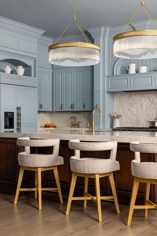A refined, traditional-style kitchen with blue-grey paneled cabinetry, dark wood island cabinetry, a pale stone benchtop and backsplash, brass fixtures, and three upholstered timber bar stools. Large tiered crystal pendant lights hang over the island, while decorative wall mouldings, arched display niches, and classical cornices add an elegant, formal character.
