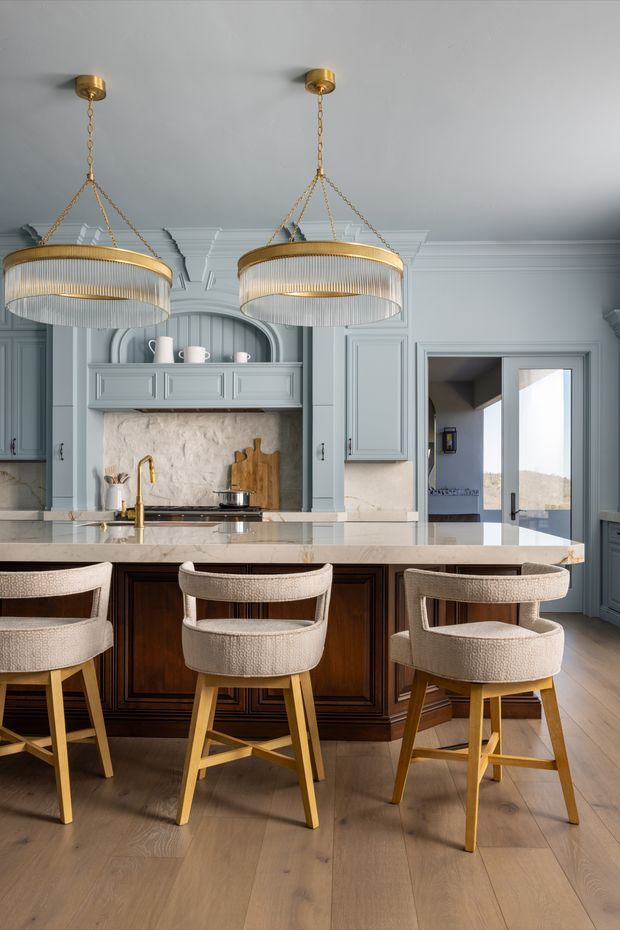 A refined kitchen interior with pale blue-grey raised-panel cabinetry, a large marble or light stone island, dark stained timber island joinery, and three upholstered bar stools. Two oversized gold-toned pendant chandeliers hang above the island. Decorative wall panelling, arched cabinetry details, warm timber flooring, and brass hardware create a polished classic look.