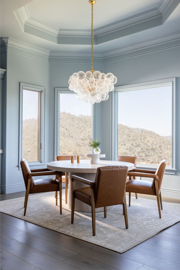 A refined dining room with a round light-wood dining table surrounded by brown leather upholstered chairs. The room features pale blue-grey walls, extensive decorative crown and wall moulding, tall framed windows overlooking a wooded hillside, dark timber flooring, and a large patterned area rug. A sculptural clustered glass chandelier hangs centrally above the table, creating a bright, elegant atmosphere.