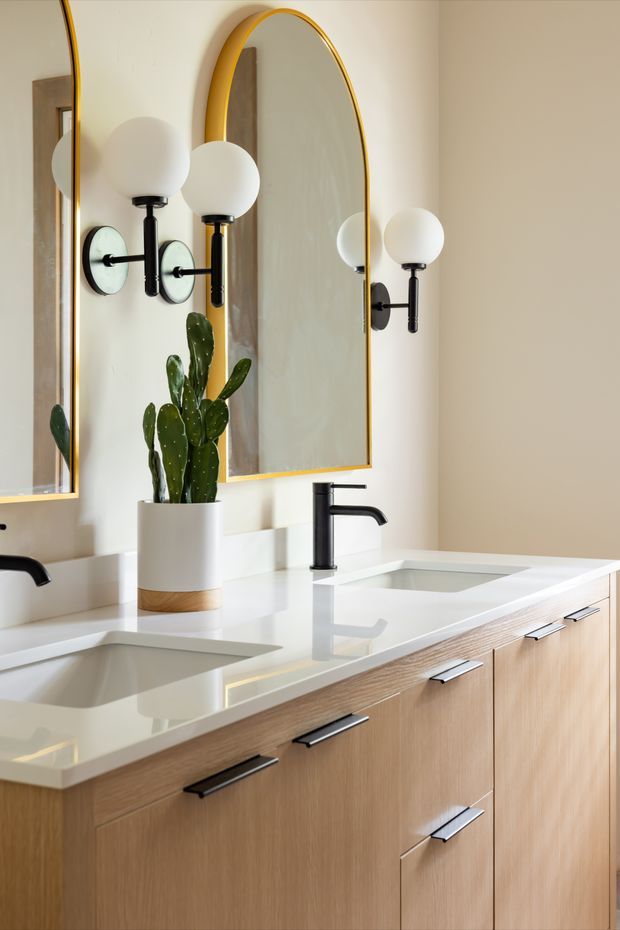 Bright contemporary bathroom vanity with a long light-wood floating cabinet, white integrated double sinks, white countertop, matte black faucets and hardware, and arched mirrors with slim brass-gold frames. Globe wall sconces flank the mirrors, while a potted cactus adds greenery. The space has a clean, refined, warm modern look.