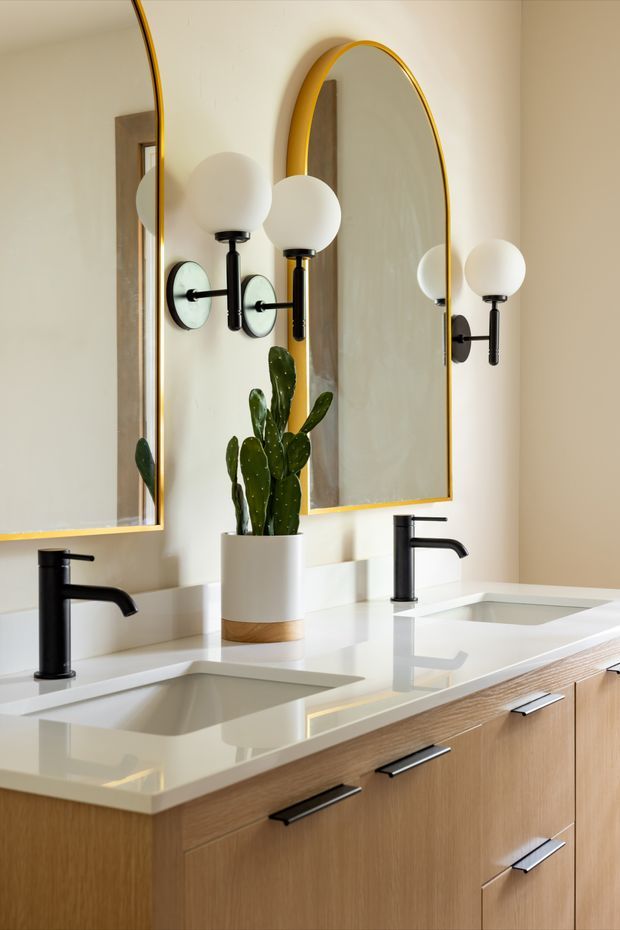 A bright contemporary double vanity bathroom with a warm light-wood floating or built-in vanity, white solid-surface countertop with two integrated rectangular basins, matte black faucets and hardware, and large arched mirrors with gold-toned frames. Globe wall sconces flank the mirrors, and a potted cactus adds greenery.