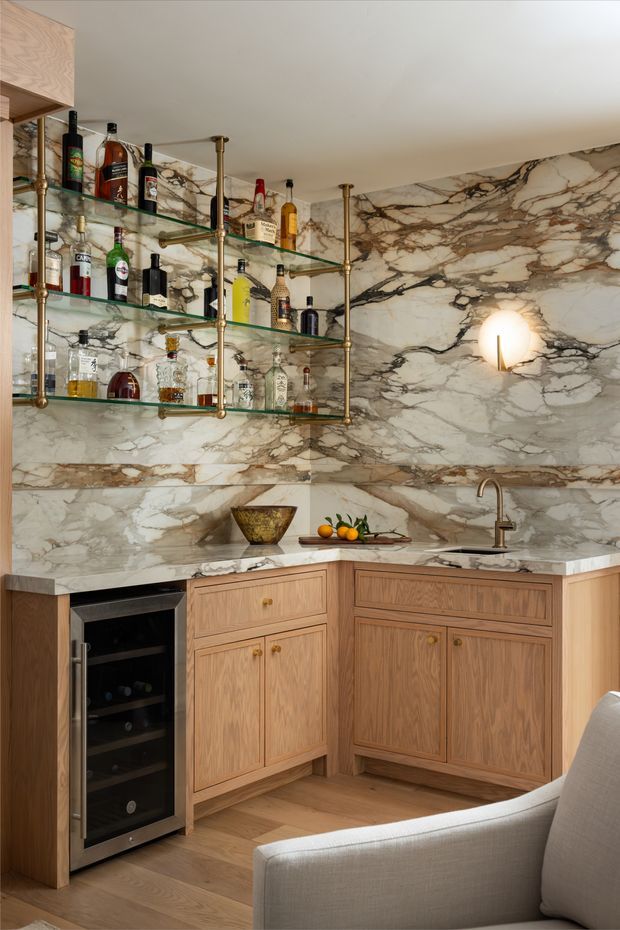 A refined home bar set within a corner of a living space, featuring full-height richly veined marble wall cladding and benchtops, warm timber cabinetry, open glass shelving supported by brass-toned metal framing, a built-in wine refrigerator, sink, and assorted bottles. A light upholstered armchair partially occupies the foreground.