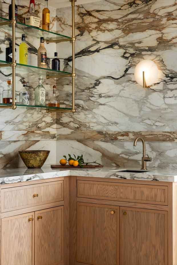 A refined home bar with a corner layout, featuring dramatic veined marble wall cladding and benchtops, warm timber cabinetry with framed fronts, brass hardware and tapware, open glass shelving supported by brass rails, and a sculptural wall sconce. Bottles and a small sink establish the bar function.