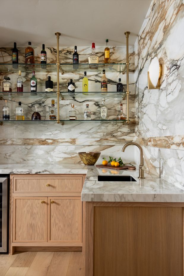 A compact, richly detailed home bar and kitchenette with full-height veined marble walls and benchtop, warm timber cabinetry, open glass shelving supported by brass uprights, a small undermount sink, and a curated selection of spirits. A brass wall sconce and warm natural timber flooring reinforce the refined, luxurious character.