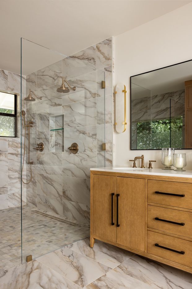 A refined bathroom featuring a large frameless glass walk-in shower, marble-look wall and floor tiles, brushed brass shower and tapware, a timber vanity with black bar pulls, a white countertop, and a large black-framed mirror. Natural light enters through a black-framed window, creating a warm, upscale atmosphere.