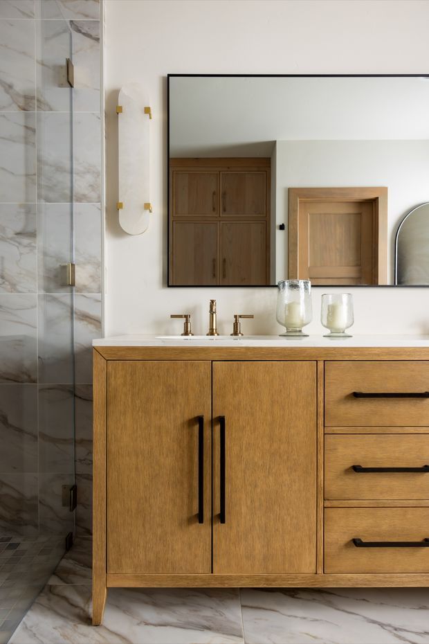 A refined bathroom vanity with warm timber cabinetry, a pale stone benchtop, brass tapware, and a large black-framed wall mirror. Veined marble-look tiles cover the shower wall and floor, while a sculptural wall light and glass vessel accessories add understated luxury.