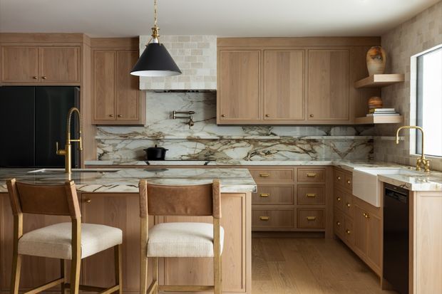 Warm, refined kitchen with natural timber cabinetry, dramatic veined stone benchtops and splashback, brass hardware and tapware, a central island with two upholstered stools, integrated dark appliances, open shelving, and a black pendant light. The space has a traditional yet contemporary feel with abundant natural light.