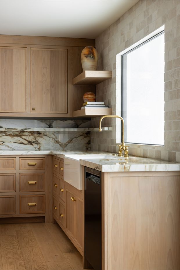 A compact, refined kitchen with warm natural timber cabinetry, brass hardware and tapware, a dramatic veined stone benchtop and backsplash, pale brick-look wall tiling, open display shelves, and a large bright window. The space has a calm, luxurious traditional-contemporary character.