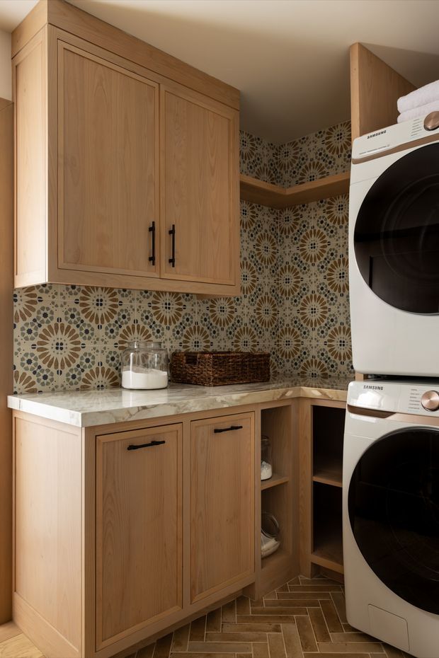 A compact, warmly lit laundry room with natural timber cabinetry, a marble-look benchtop, patterned floral geometric tiled backsplash, open shelving, and a stacked white front-loading washer and dryer. The floor is laid in a herringbone pattern, and woven storage adds texture.