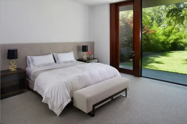 A bright, contemporary bedroom with a low upholstered bed and textured beige headboard, white bedding, matching bedside tables and black-and-gold lamps. A long upholstered bench sits at the foot of the bed. Floor-to-ceiling timber-framed glazing opens the room visually to a green garden, while the carpeted floor and restrained palette create a calm, refined atmosphere.