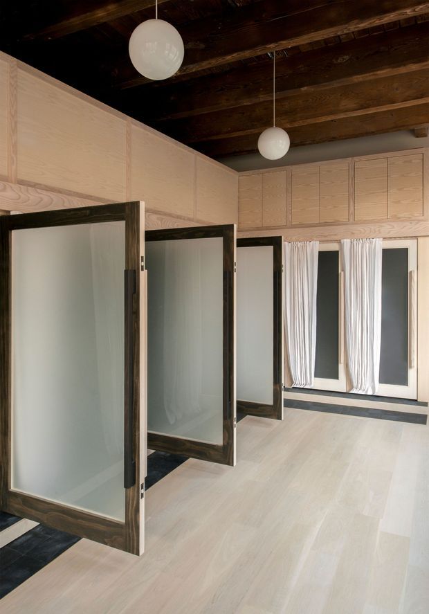 A refined interior corridor or changing area with a series of dark timber-framed frosted-glass doors, pale timber wall panelling and cabinetry, and a light timber floor. The space has exposed dark wood ceiling beams, sheer white curtains, dark inset floor mats, and simple spherical pendant lights.