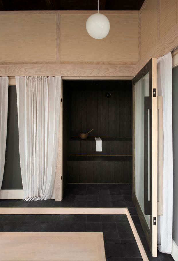 A refined compact interior clad in pale timber, featuring a dark tiled floor and a recessed dark timber-lined bathing or shower alcove with built-in shelves. Sheer white curtains, black-framed glazing, and a spherical pendant light create a calm, minimalist contrast between light wood and charcoal surfaces.