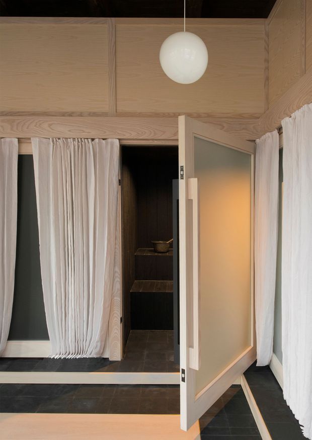 A refined interior transition space with a dark tiled floor, pale timber wall and ceiling panelling, sheer white curtains, and a large frosted-glass timber door opening into a compact dark-lined room with stepped platforms. A simple white globe pendant adds soft illumination, while warm light glows through the frosted glass.