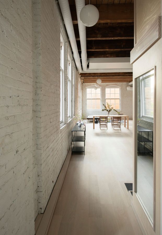 A long, bright loft-style interior with white-painted exposed brick walls, pale timber flooring, tall industrial windows, exposed dark timber ceiling beams, and visible white ductwork. A simple timber dining table and chairs sit at the far end, with a narrow black console along the wall and a large reflective glazed panel on the right. Globe pendant lights and restrained furnishings create an airy, refined atmosphere.
