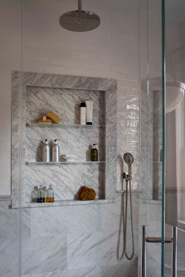 Close-up view of a refined walk-in shower with white glossy subway wall tiles, large marble-look wall tiles, and a recessed marble-lined niche with glass shelves holding toiletries. Brushed metal rain shower and handheld shower fittings are visible behind a clear glass shower screen and door.