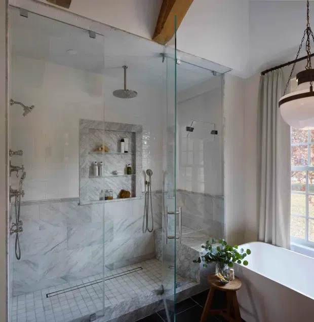 Bright, refined bathroom with a large frameless glass walk-in shower, dual shower fittings, marble-look wall cladding, recessed shelving, and small mosaic shower-floor tiles. A freestanding white bathtub sits beside a tall gridded window with soft curtains. Exposed timber ceiling beams and a suspended pendant add warmth to the otherwise light, neutral space.
