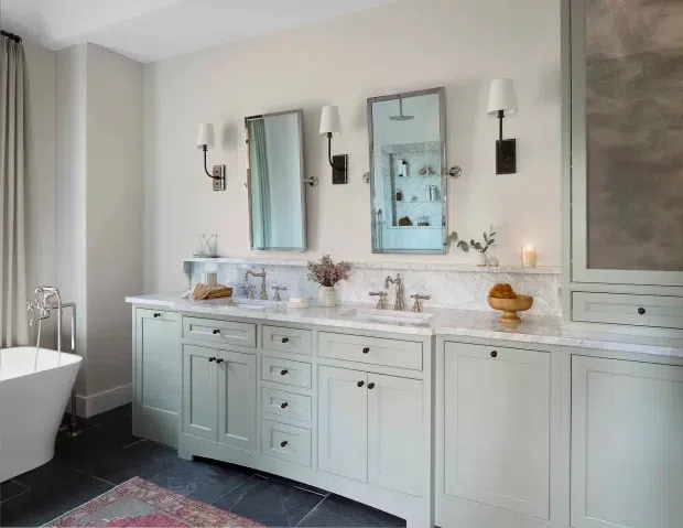 Elegant traditional bathroom with a long pale painted double vanity, marble countertop and backsplash, twin undermount sinks, polished metal fittings, framed mirrors, wall sconces, freestanding bathtub, dark tile flooring, and built-in cabinetry. The room has a refined, softly lit, classic appearance.
