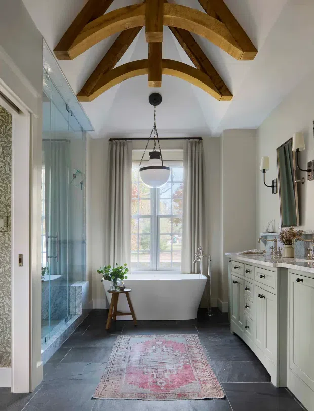 A bright, refined bathroom with a freestanding white bathtub positioned beneath a large window, a glass-enclosed walk-in shower, and a long pale built-in vanity with dark hardware and a stone countertop. The room features dark slate-like flooring, pale walls, tall curtains, wall-mounted sconces, and a dramatic vaulted ceiling with exposed curved timber beams. A pink patterned runner and small plant add warmth and colour.