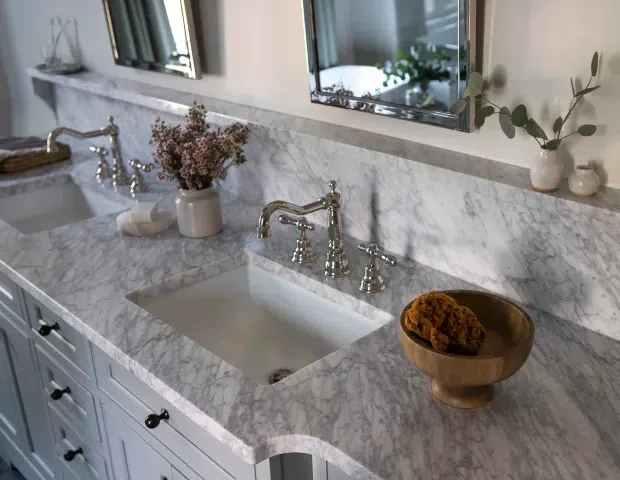 A refined bathroom vanity with twin undermount basins set into a continuous white-and-grey veined stone benchtop and matching splashback. Polished chrome bridge-style tapware, pale blue-grey shaker cabinetry with dark knobs, framed mirrors, and restrained botanical and ceramic accessories create a classic, elegant look.