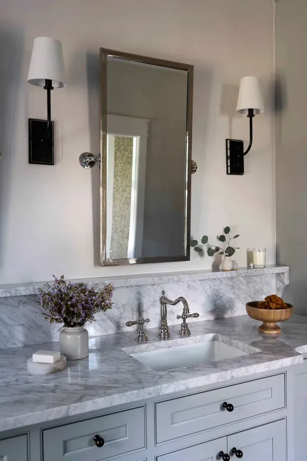 A refined bathroom vanity vignette featuring a large rectangular framed mirror, symmetrical wall sconces, a veined marble countertop and backsplash, an undermount rectangular sink, polished metal bridge-style tapware, and pale blue-grey shaker cabinetry. Small decorative plants, flowers, and a bowl add warmth to the otherwise classic composition.