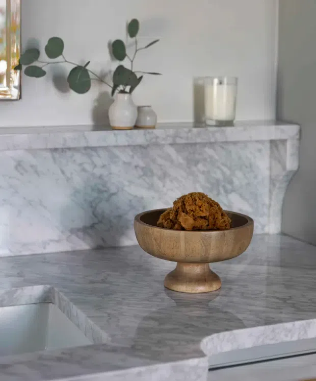 Close-up interior detail of a refined marble fireplace surround and hearth, with a veined white-grey marble mantel and ledge. A wooden pedestal bowl, candle, ceramic vessels, and eucalyptus branches provide understated styling against a pale neutral wall. The image is softly lit with a calm, elegant atmosphere.