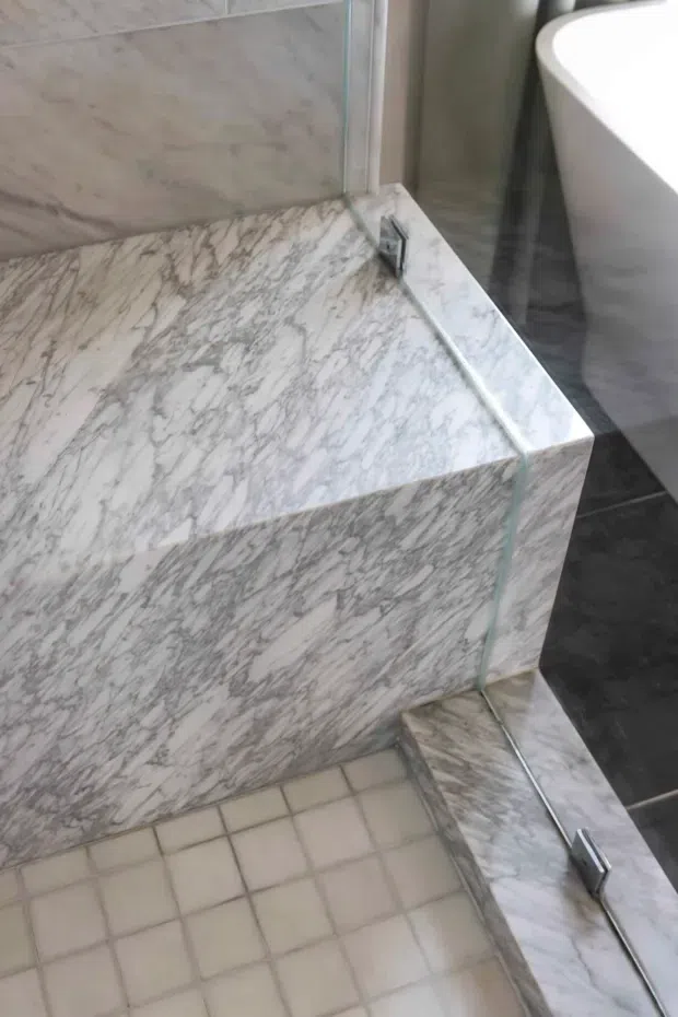Close-up view of a bathroom featuring a marble-lined raised bath or shower platform, glass shower screen with metal fittings, small square floor tiles, and a partially visible white freestanding bathtub.