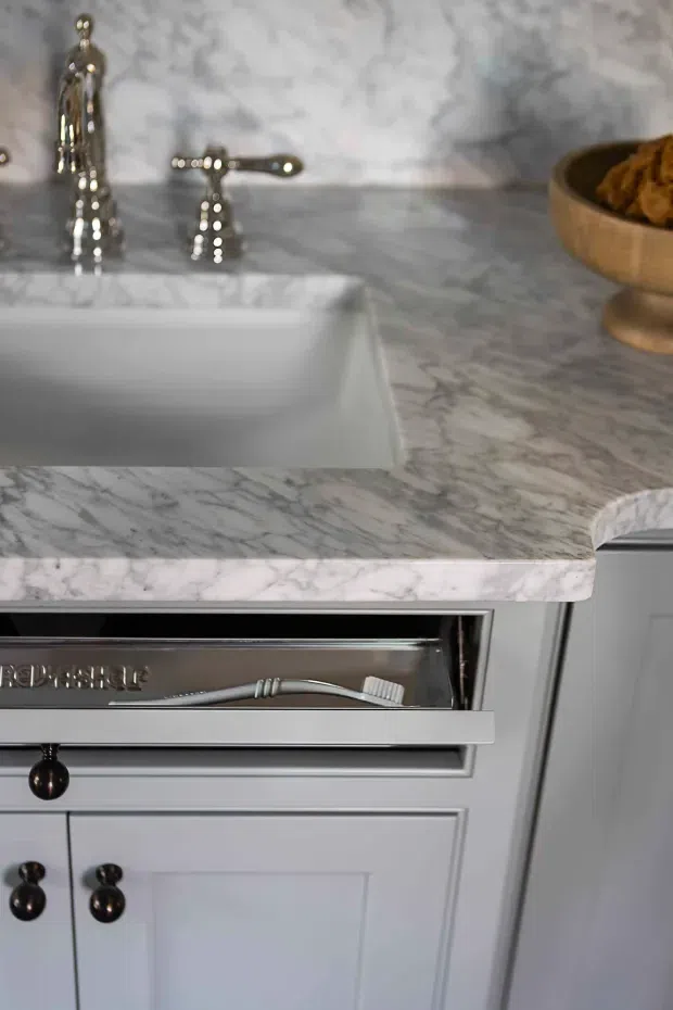 Close-up of a refined bathroom vanity with a white marble countertop and integrated rectangular basin. Chrome traditional-style tapware sits against a marble backsplash, while painted grey-blue shaker cabinetry with dark round hardware includes an open drawer containing grooming items. A wooden bowl provides a warm accent.