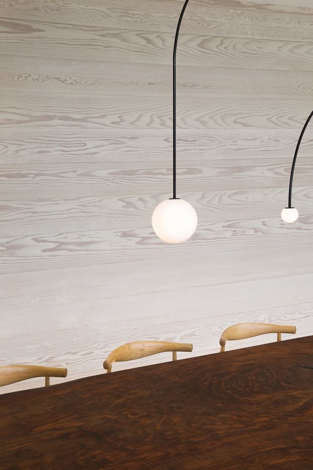 Close-up interior view of a refined dining area, featuring a pale horizontal timber wall lining with prominent natural grain, two simple spherical pendant lights on black cords, and the upper portions of curved timber dining chairs around a dark richly grained timber dining table. The composition is minimalist and material-focused, with warm wood contrasting against the light wall finish.