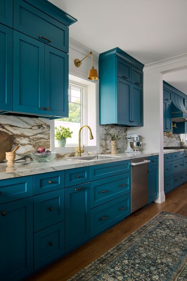 A richly detailed kitchen with deep teal-blue Shaker-style cabinetry, ornate crown moulding, polished marble benchtops and matching marble splashback. Brass hardware, a brass gooseneck tap, under-mount sink and brass wall sconce add warmth. A stainless-steel dishwasher, stand mixer, patterned rug and timber flooring complete the traditionally styled space. A second section of the kitchen is visible through the opening at right.