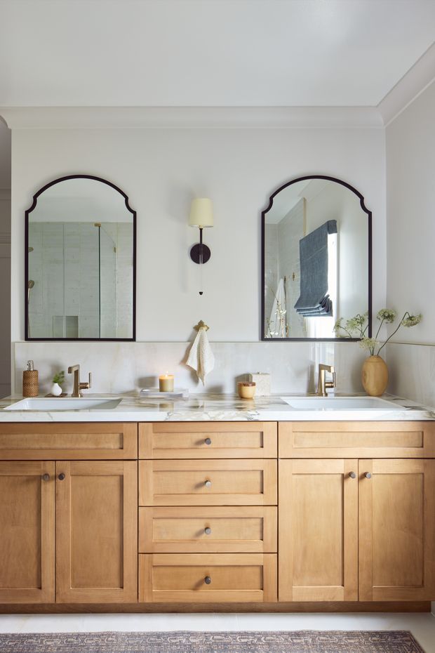 A bright, refined bathroom vanity with a wide double-basin timber cabinet, white marble countertop and backsplash, brass-toned faucets, and two arched dark-framed mirrors. Classic wall mouldings, a central wall sconce, and simple ceramic and botanical accents create a warm, traditional feel.