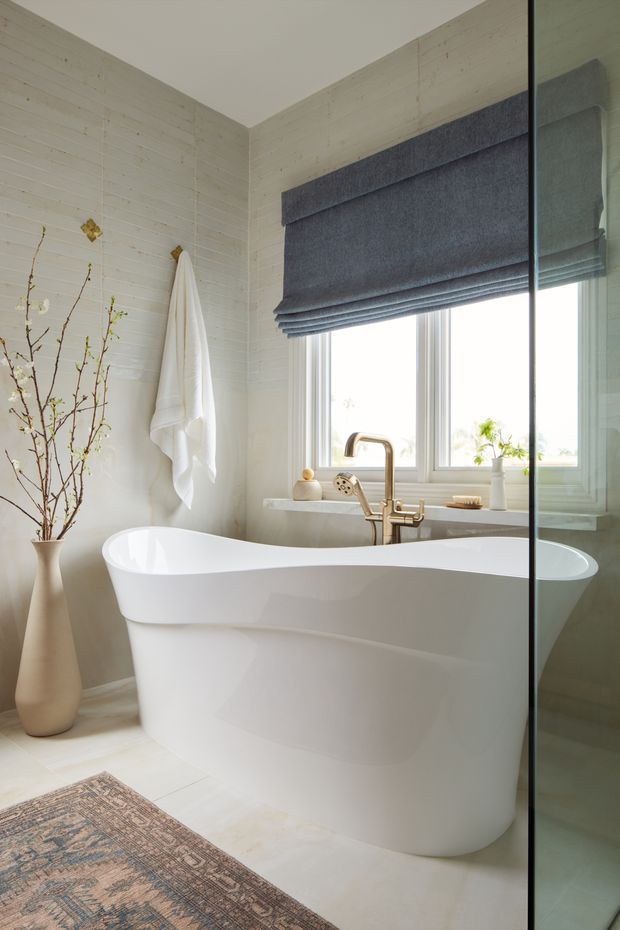 Bright, refined bathroom featuring a freestanding white oval bathtub positioned beneath a window, with brushed metal tapware, a grey Roman blind, pale horizontal wall panelling, light flooring, and a glass shower enclosure. Natural branches in a vase and a patterned rug add organic and muted decorative accents.