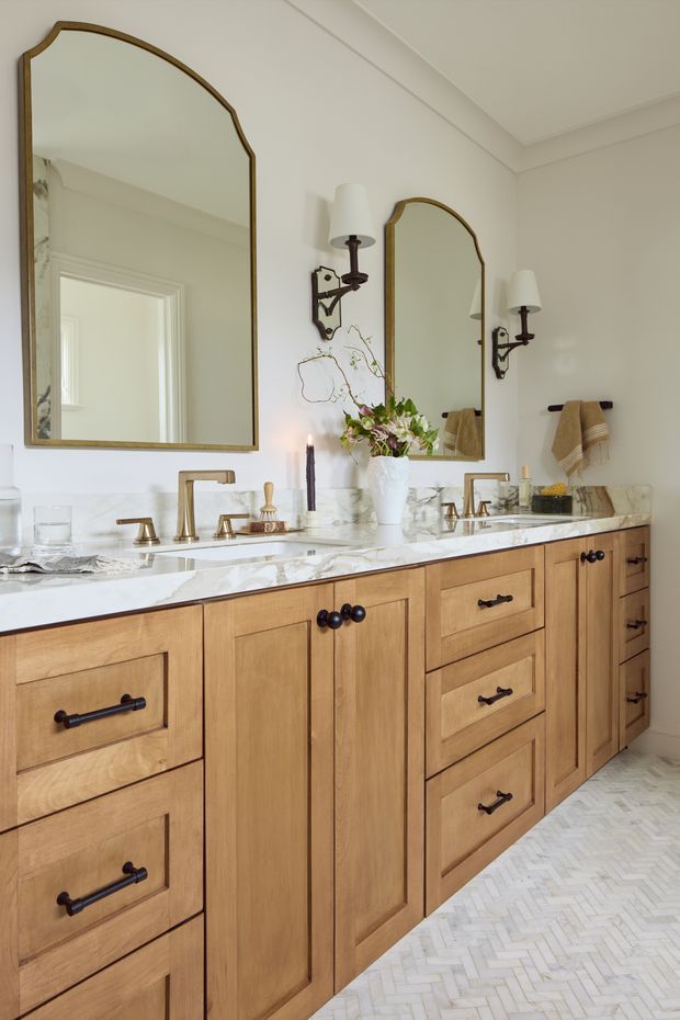 Elegant double-vanity bathroom with warm natural wood cabinetry, a white marble countertop and backsplash, polished brass tapware, ornate brass-framed mirrors, wall sconces, and a light herringbone tile floor. The room has a refined traditional aesthetic with layered decorative detailing.