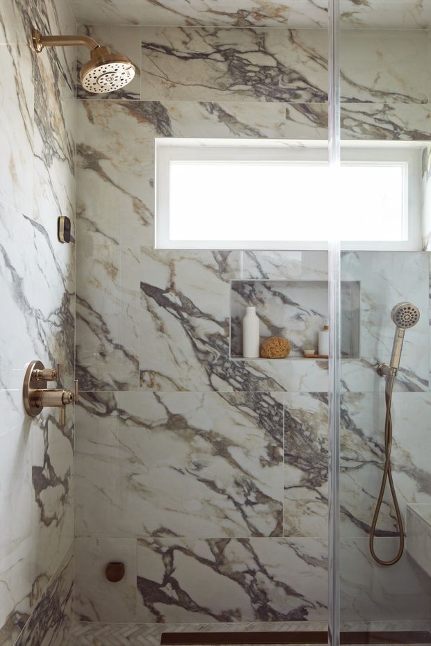 A compact, luxurious walk-in shower enclosed by clear glass, with dramatic veined natural stone or marble-look wall and ceiling tiles. Brass shower fixtures, a handheld shower, recessed niche, small horizontal window, and patterned floor create a refined contemporary bathroom interior.
