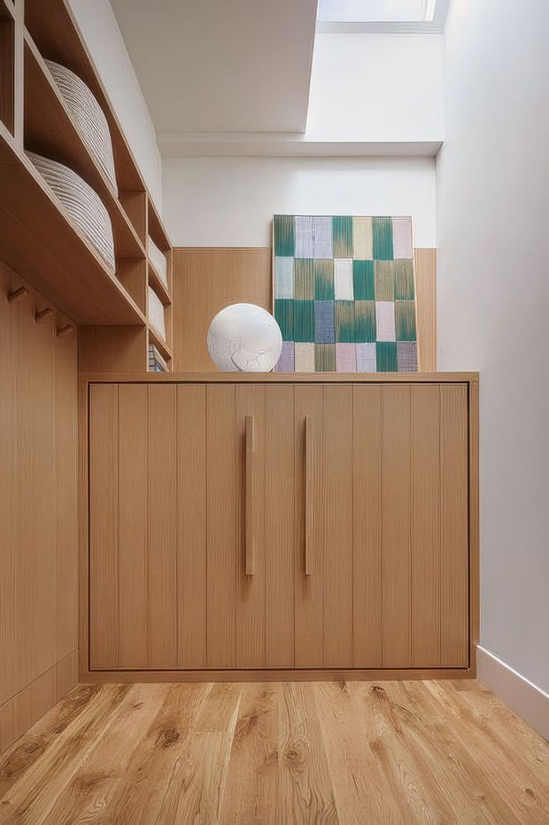 A compact, softly lit built-in storage area with warm light-wood cabinetry, vertical fluted detailing, long integrated handles, open shelving, and a matching timber floor. A skylight brings daylight into the white-walled space, while a colourful geometric artwork and ceramic object add restrained decorative accents.