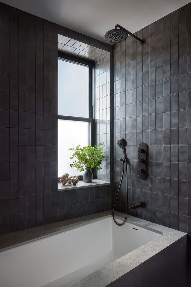 A moody, contemporary bathroom featuring a built-in white bathtub beneath a tall frosted window. Dark charcoal square wall tiles continue around the shower and bath, with contrasting lighter grey tile detailing at the window reveal and tub surround. Matte black shower fixtures include an overhead rain shower, handheld shower, controls and bath spout. A small plant and decorative objects sit on the deep window sill.