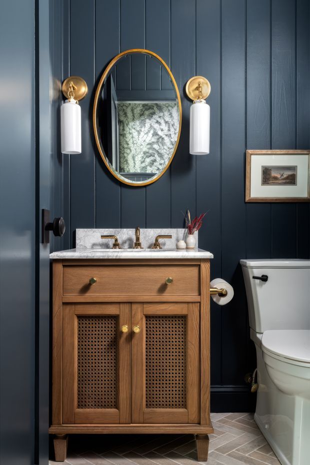 A compact, richly styled bathroom featuring a warm timber vanity with cane-front cabinet doors, a pale stone benchtop and brass tapware. Dark navy vertical wall panelling creates a dramatic backdrop, complemented by an oval brass-framed mirror and matching wall sconces with white cylindrical shades. A toilet and framed artwork are visible, with patterned grey-brown floor tiles.