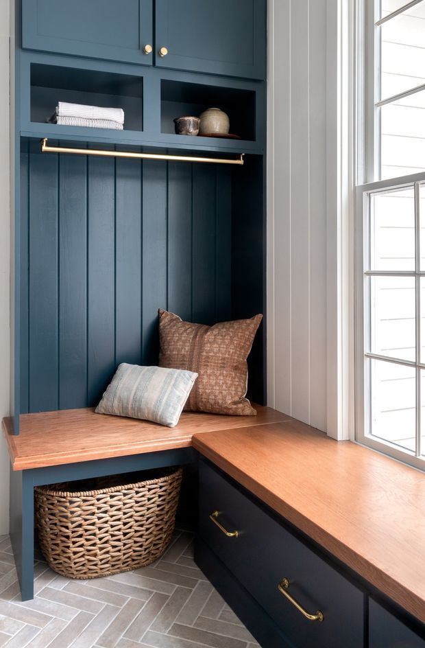 A compact built-in entryway or mudroom nook beside a large divided-light window. Deep navy painted cabinetry and vertical wall panelling frame open display shelves, a timber bench seat with cushions, a brass hanging rail, and lower drawers. A woven basket sits beneath the bench, while warm timber countertops and grey patterned floor tiles add contrast. The space is bright, tailored, and functional with classic detailing.